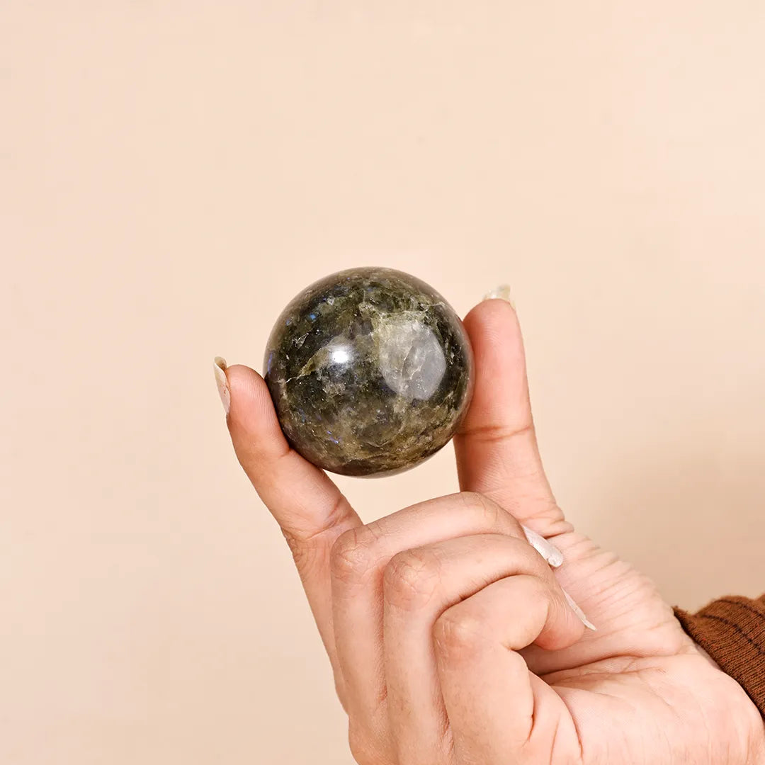Hand holding a polished stone sphere against a beige backgroundNatural Labradorite Crystal Ball – Abhimantrit & Certified