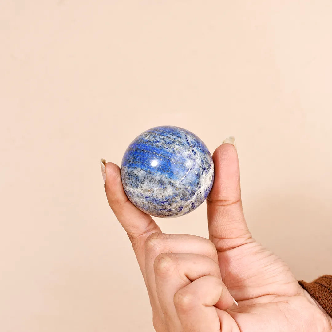 Hand holding a blue and white marbled sphere against a beige backgroundNatural Lapis Lazuli Crystal Ball – Abhimantrit & Certified