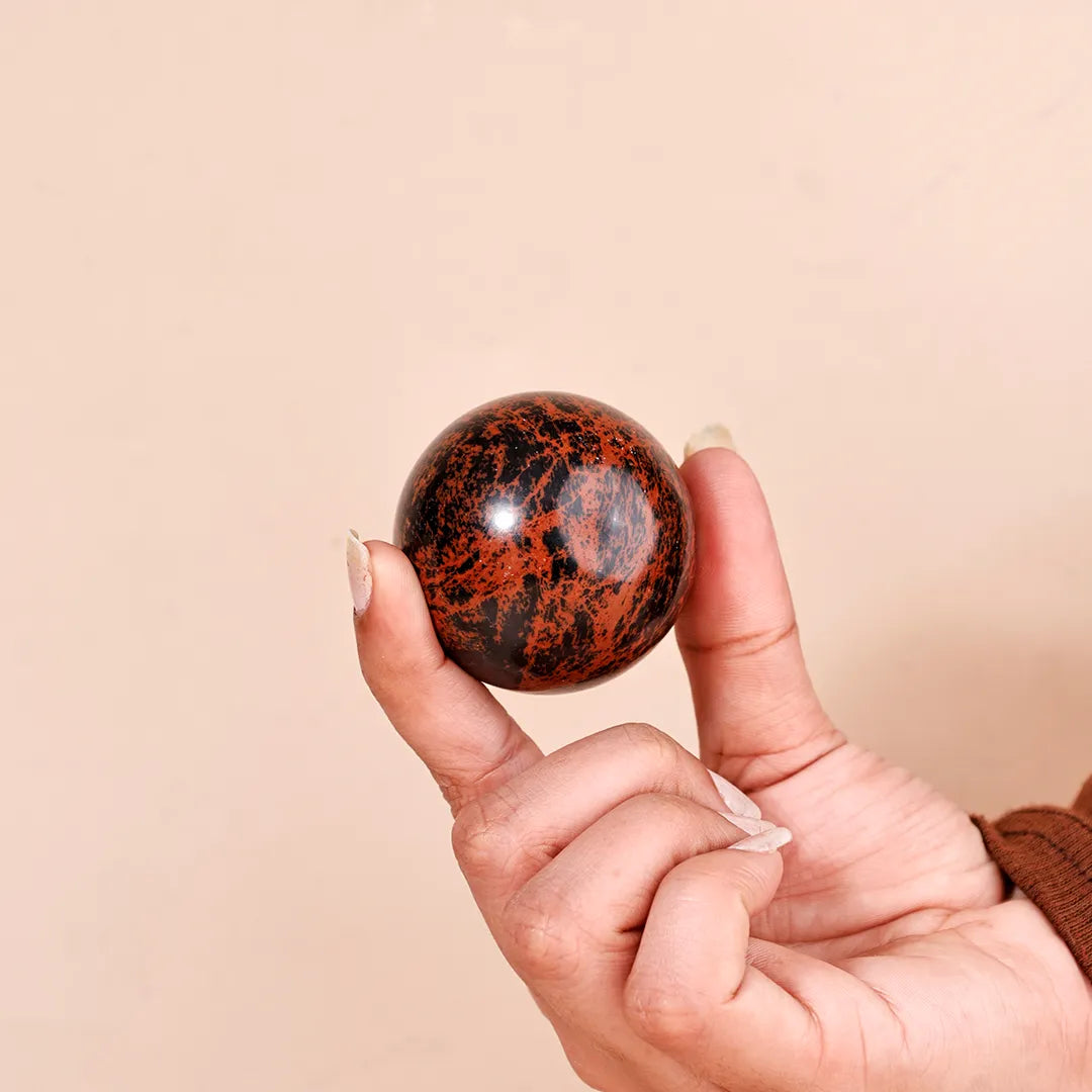 Hand holding a marbled stone ball against a beige backgroundNatural Mahogany Obsidian Crystal Ball – Abhimantrit & Certified