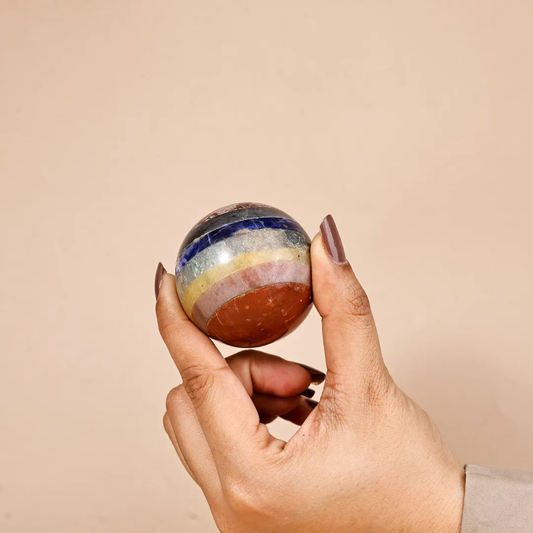Hand holding a multicolored spherical object against a beige backgroundNatural 7 Chakra Crystal Ball – Abhimantrit & Certified