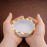 Hands holding a small white dish with gold decorative edge on a brown background