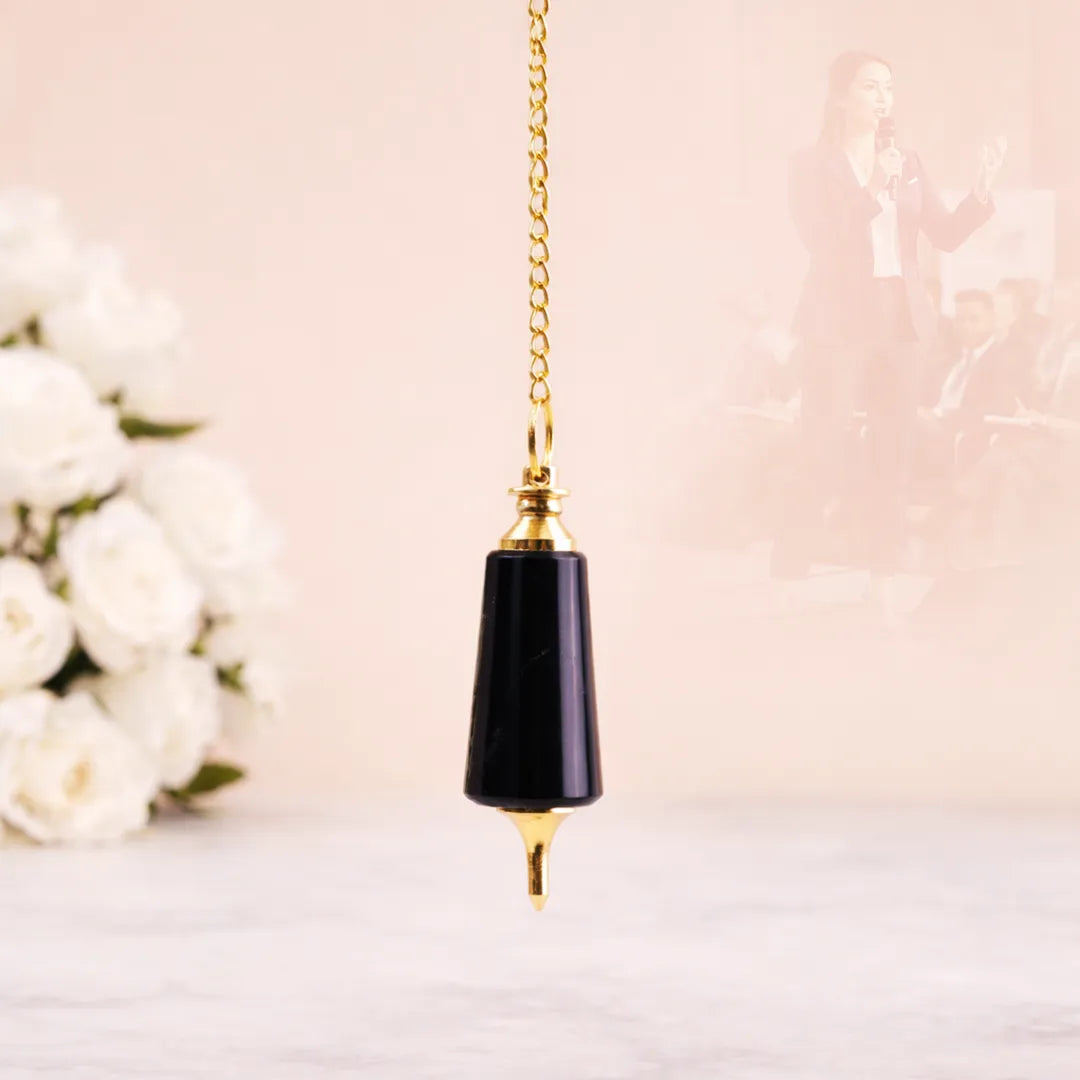 Decorative black and gold pendant on a chain against a soft pink background with white flowers.