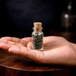 Hand holding a small glass bottle with cork lid containing green stones against a dark background