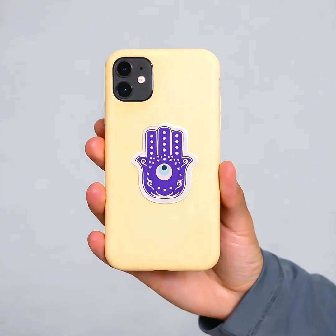 Hand holding a yellow phone case with a purple Hamsa hand design on a gray background