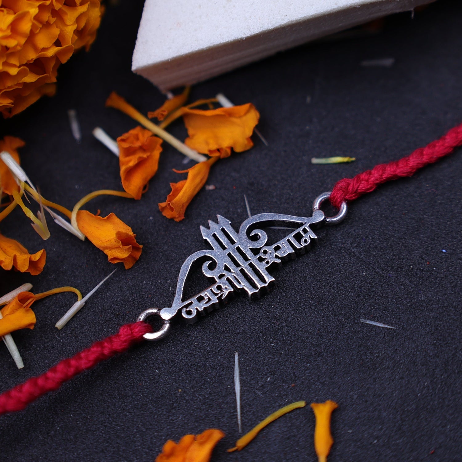 A red string Rakhi bracelet with a silver-colored jai shree shyam charm, styled on a black surface with orange marigold flowers and a white box.