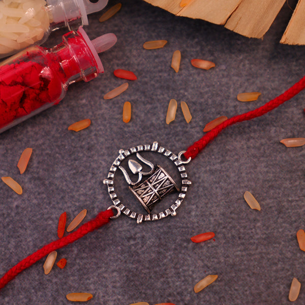Handcrafted 925 silver Damru and Trishul Rakhi with red adjustable thread, inspired by Lord Shiva, displayed on a decorative background with kumkum, rice grains, and wooden accents. Traditional Raksha Bandhan bracelet, spiritual Hindu gift for brother, sacred Shiva rakhi for festival celebration.