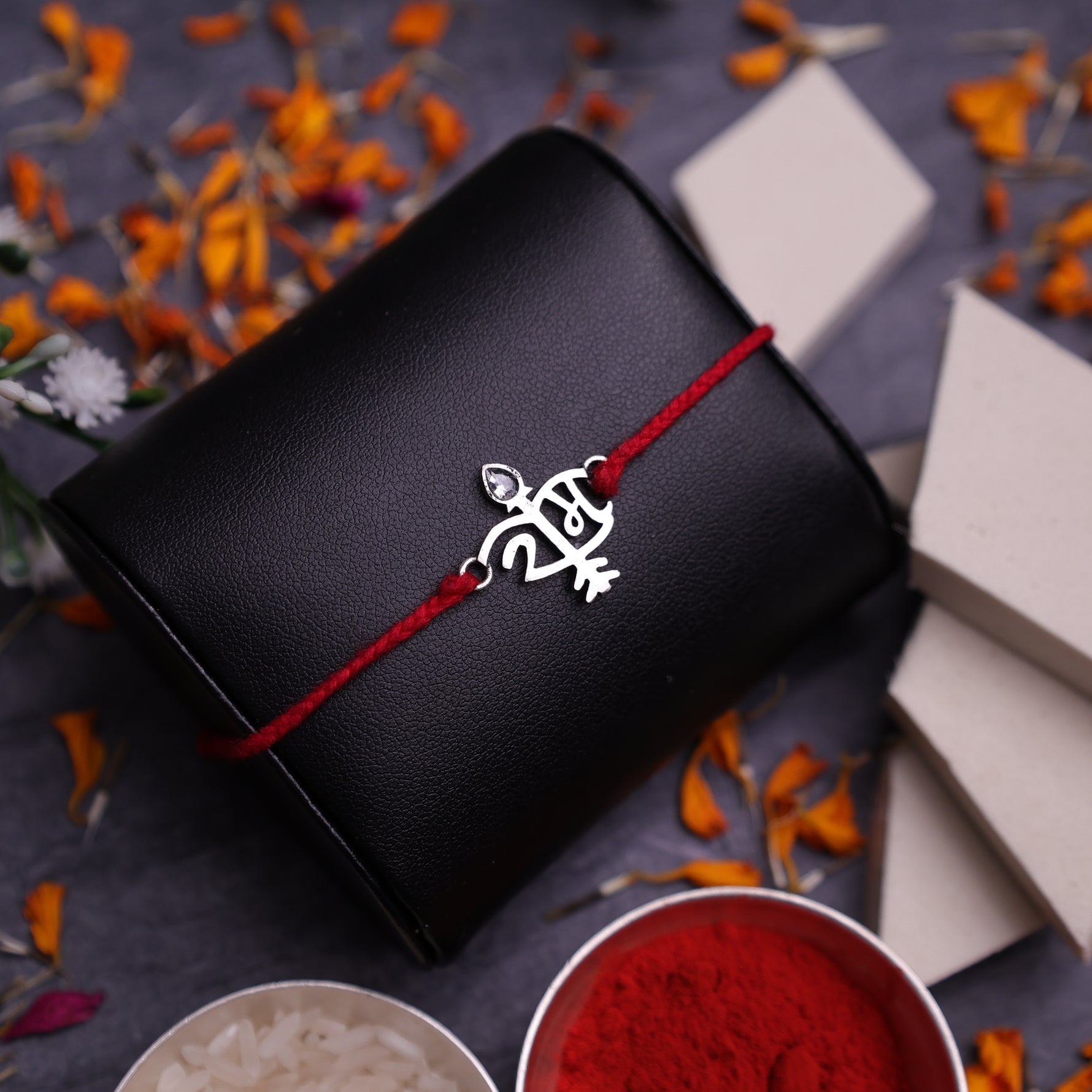 A Rakhi bracelet with a 925 silver Ram symbol and red thread, presented on a dark Black background with decorative orange flower petals, sweets, and bowls of powder.