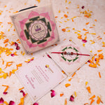 A boxed Rakhi gift set with a pink and green lotus-patterned box, a red beaded Rudraksha Rakhi with 925 silver OM Symbol with a red thread The box and cards show the brand name Dhwani Astro.