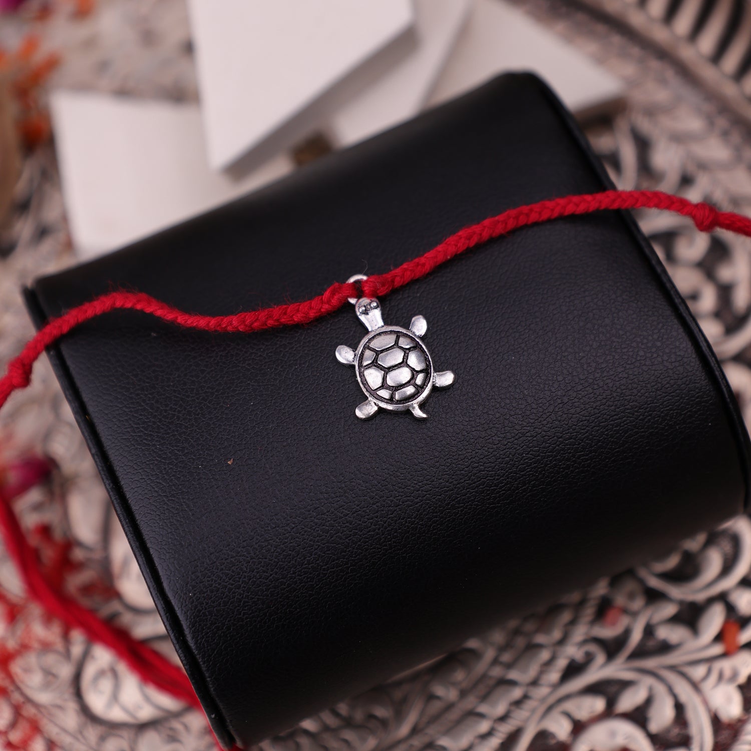 A rakhi bracelet with a small silver turtle charm, displayed across a black textured cylindrical holder on a patterned surface.