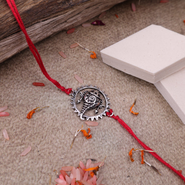 Handcrafted 925 silver conch shell rakhi  with a vibrant red thread, displayed on a neutral stone-textured background with decorative flower petals, wooden log, white display blocks