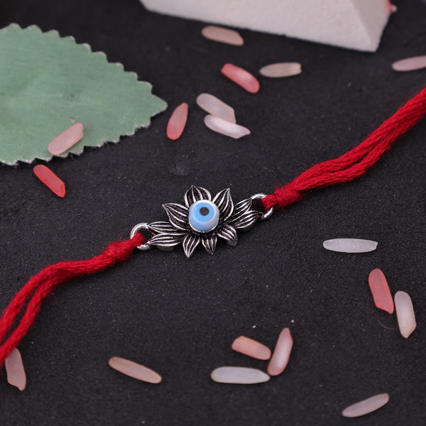 A red string bracelet with a silver-toned lotus flower charm featuring a light blue evil eye bead, displayed on a dark textured surface with scattered pink and white beads.