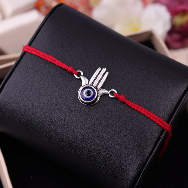 A silver Hamsa hand evil eye charm on a red cord bracelet, presented on a black textured pouch inside a gift box.