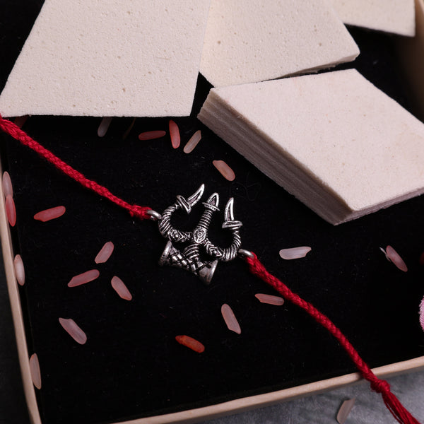 Handcrafted silver-tone Trishul and Damru Rakhi with an adjustable red thread, inspired by Lord Shiva, displayed on a neutral gray background with decorative rice grains and white stone props. Traditional Hindu Raksha Bandhan bracelet, spiritual brother gift, Shiva Trident Rakhi for festive celebration and devotional wear.