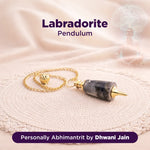 Labradorite pendulum with gold chain on a soft fabric background, branded as 'Personally Abhimantrit by Dhwani Jain'.