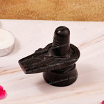 A black stone Shiva Lingam idol on a light marble surface with a textured gold background, featuring a candle holder and flower petals in the periphery.