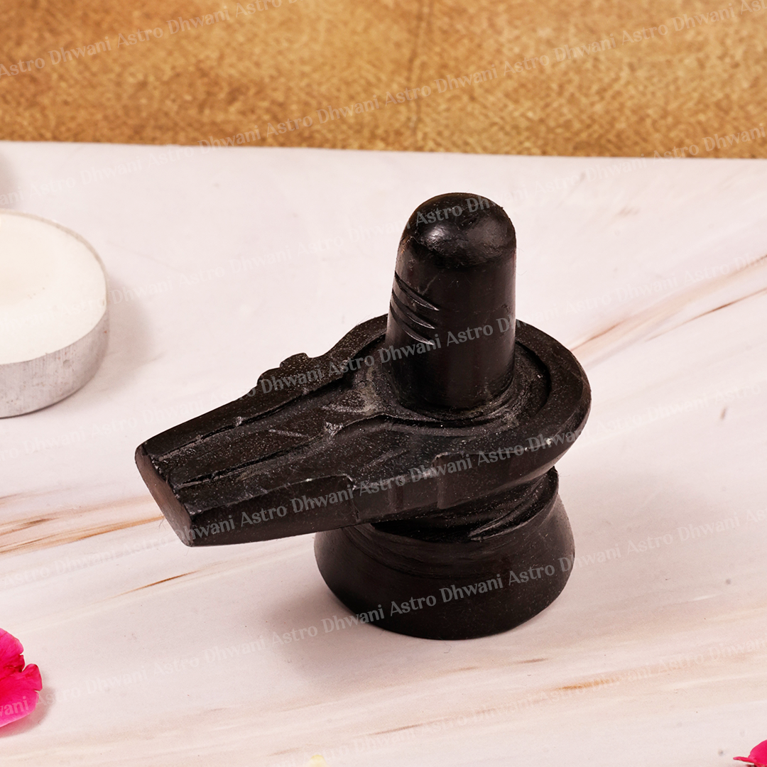 A black stone Shiva Lingam idol on a light marble surface with a textured gold background, featuring a candle holder and flower petals in the periphery.
