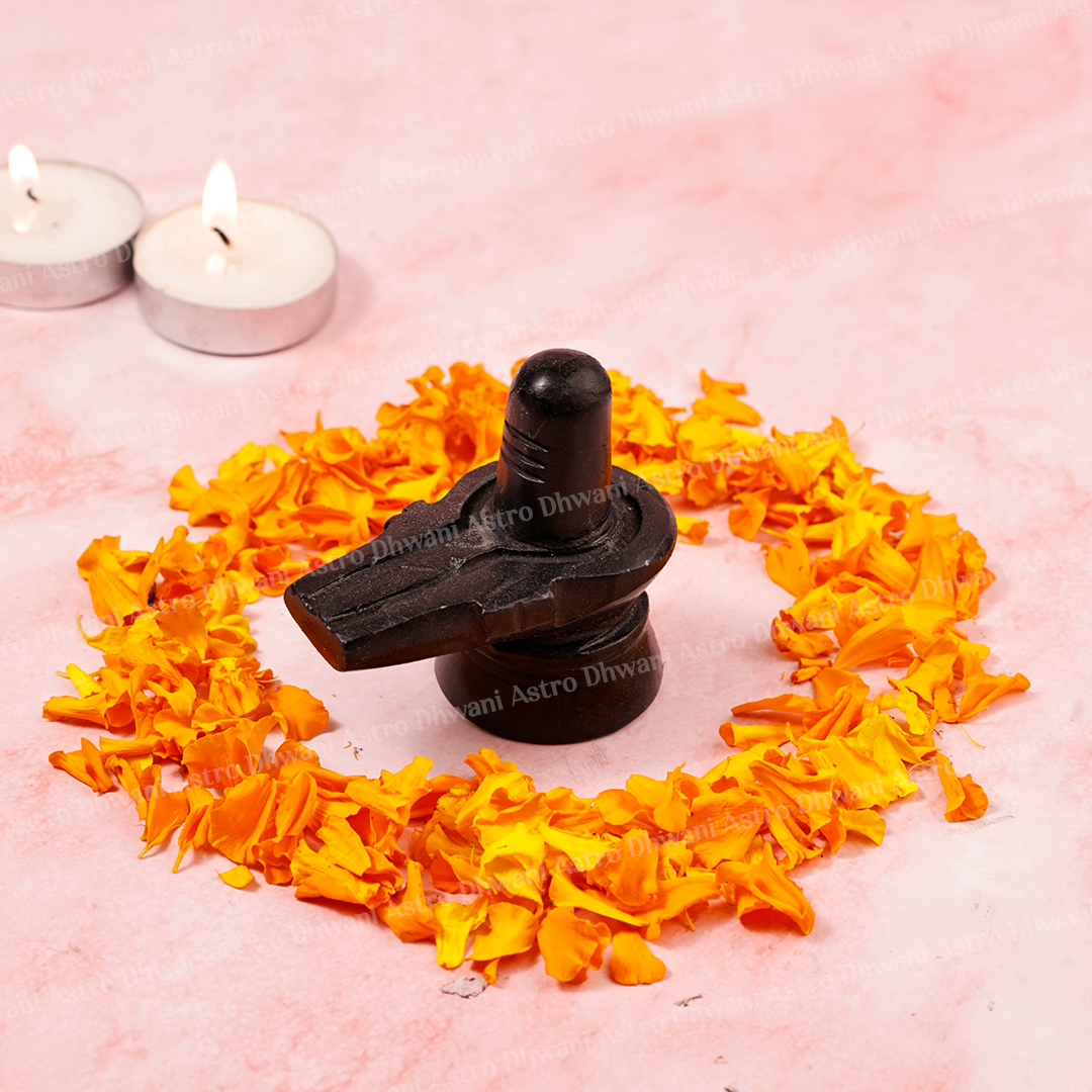 A black stone Shiv Lingam placed on a pink surface, surrounded by a circular arrangement of orange marigold petals, with two lit tealight candles in the background.Natural Black Marble Shivling | Handcrafted Shiva Lingam for Home Temple, Puja & Meditation | Sacred Marble Shivling Idol