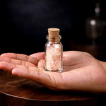 Hand holding a small glass bottle with pink Himalayan salt on a wooden surface.
