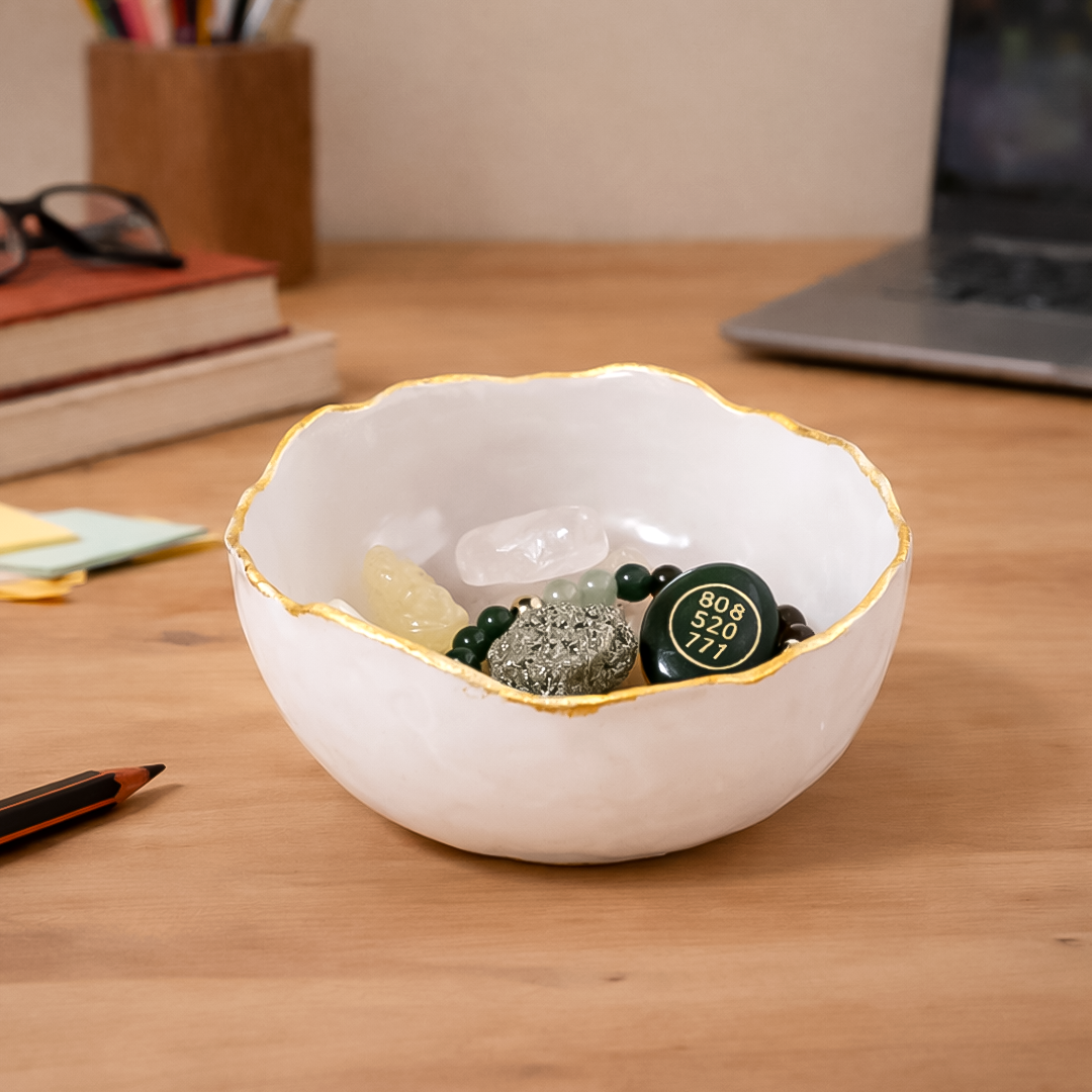 White decorative bowl with gold rim on a wooden desk containing small stones and a round object.