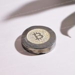 A circular metallic Bitcoin token or coin featuring a gold-toned center with a black Bitcoin symbol, photographed on a white surface with a strong shadow.