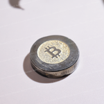 A round, metallic cryptocurrency token featuring a gold Bitcoin symbol in the center, set against a plain white background with a soft shadow.