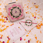 A boxed Diwali gift set arranged on a light textured surface with scattered orange and pink flower petals. The set includes a pink lotus-themed box labeled 'Diwali Gift Set' with a red and silver beaded rakhi laid across one card, plus a pink greeting card.