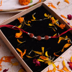 A red cord bracelet with three brown rudraksha beads and silver spacer beads, presented in a beige gift box with a black velvet lining and scattered orange flower petals.