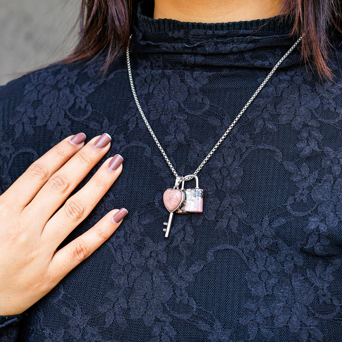 Person wearing a dark lace top with a silver necklace featuring a heart and key pendant.